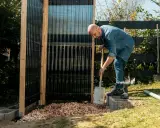 Uomo costruisce una doccia da giardino fai da te, livellando ghiaia con una pala.