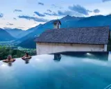 Due donne in una piscina a sfioro ammirano il panorama di Bormio e dintorni, con montagne maestose e un piccolo edificio storico.