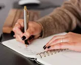 Dłoń z ciemnym lakierem pisze w notatniku. Może to lista zadań, a może spis potencjalnych poręczycieli dla kredytu?