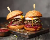 Dwa soczyste burgery z grilla, z jajkiem sadzonym i dodatkami, prezentują się apetycznie na desce.