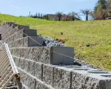 Budowa muru oporowego z bloczków betonowych. Widać drabinę i różową nić, która wyznacza poziom. Odległość od granicy działki jest kluczowa dla prawidłowego posadowienia.