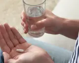 Dłoń trzymająca białą kapsułkę i szklankę wody.