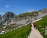 Una persona con zaino e bastoncini da trekking percorre un sentiero fiorito durante un'escursione sul Gran Sasso, con montagne imponenti sullo sfondo.