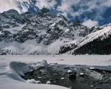 Morskie Oko zimową porą. Lodowate wody jeziora odbijają ośnieżone szczyty Tatr, a na horyzoncie widać ludzi spacerujących po zamarzniętej tafli.