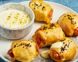 Goldbraune Blätterteigkissen mit Würstchen, bestreut mit schwarzen Sesamkörnern, serviert mit einer cremigen Dip-Sauce.