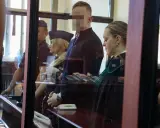 Osoba w kajdankach, prawniczka i policjanci w sali sądowej.