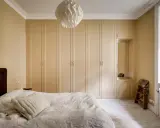 Sovrum med en stor, ljusbeige garderob som sträcker sig längs väggen. En vit taklampa med geometriskt mönster hänger ovanför.