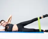 Femme souriante faisant des exercices de jambes avec une bande élastique, travaillant les fentes latérales sur un tapis bleu.