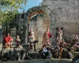 Un groupe de guerriers, prêts à combattre, pose devant des ruines antiques. Ces hommes, tels des gladiateurs romains, arborent armures et armes, prêts pour l'arène.