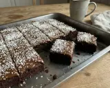 En kladdkaka med glasyr och kokosflingor skuren i bitar i en plåt. En mugg kaffe står bredvid.