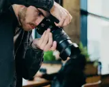 Mężczyzna z brodą w czarnej kurtce trzyma aparat, gotowy do uchwycenia chwili w stylu street photography.