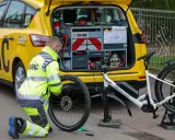 ADAC-Mitarbeiter repariert eine Fahrradpanne. Ein gelbes ADAC-Auto mit Werkzeugkasten steht daneben.