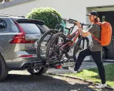 Mężczyzna z pomarańczowym plecakiem montuje bagażnik rowerowy na hak do Volvo XC60. Dwa rowery górskie są już zamocowane. Przepisy dotyczące bagażnika rowerowego na hak są kluczowe dla bezpieczeństwa.