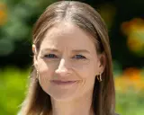 Porträt einer Frau mit blauen Augen und braunem Haar, das einige einzelne graue Haare abdeckt. Sie trägt goldene Ohrringe.