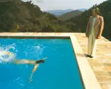 A portrait of an artist by a pool with two figures. One figure swims in the bright blue water, while another stands on the tiled edge, looking out at the mountainous landscape.
