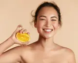 Donna sorridente con fetta d'arancia, suggerisce che il siero alla vitamina C si può usare in estate per una pelle luminosa.