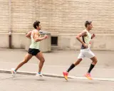 Zwei Läufer auf einer Straße, die versuchen, ihren **pace verbessern**. Der vordere Läufer trägt ein hellgrünes Tanktop und schwarze Shorts, der hintere ein gelb-graues Tanktop und weiße Shorts.