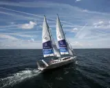 Le voilier Pangaea de Mike Horn navigue sur une mer calme sous un ciel bleu parsemé de nuages et de traînées d'avions.