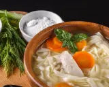 Rosół z makaronem, kawałkami kurczaka i marchewką. Obok miseczki z posiekanym szczypiorkiem, koperek i sól, która wzmacnia smak, jak glutaminian.