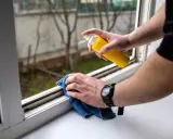 Persona rocía un limpiador en un marco de ventana sucio y usa un paño azul para limpiar moho y suciedad.