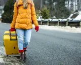 Kobieta w żółtej kurtce i czerwonych rękawiczkach ciągnie żółtą walizkę. W tle zimowy krajobraz. Modlitwa o szczęśliwą podróż.