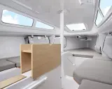 Intérieur du FIRST 24, cabine lumineuse avec banquettes grises, table en bois et hublots ouverts sur le ciel bleu.