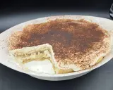 Un délicieux tiramisu sans oeuf, crémeux et saupoudré de cacao, dans un plat blanc. Une part a été retirée, révélant les couches de biscuits et de crème.