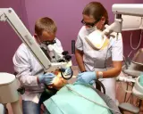 Dwaj dentyści w białych fartuchach pracują przy pacjencie, używając mikroskopu stomatologicznego.