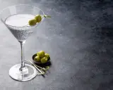 Ein Glas trockener Wermut mit Oliven garniert, daneben eine Schale mit weiteren Oliven auf dunklem Untergrund.