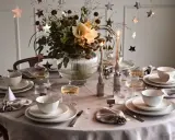 Enkel nyårsdukning med vita tallrikar, champagneglas, stjärnformade dekorationer och en blomsterbukett i en glaskaraff.