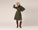 Elegancki, oliwkowy rodzaj płaszcza z wiązaniem w talii i kontrastowymi szwami. Modelka prezentuje go z wysokimi, brązowymi kozakami.
