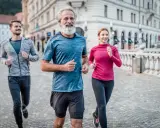 Un uomo con barba bianca e due giovani corrono sorridenti in città. Un esempio di come iniziare a correre a 50 anni sia possibile e gratificante.