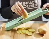 Eine Hand schneidet Kartoffeln für ein Kartoffelsandwich auf einem Hobel. Dünne Scheiben liegen auf dem Holzbrett.