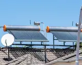 Dos calentadores de agua caliente solar en un tejado, junto a antenas parabólicas, bajo un cielo azul.