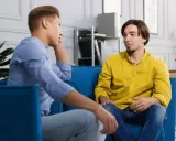 Dwóch mężczyzn siedzi na kanapie, rozmawiając. Jeden z nich opowiada o tym, jak przełamać barierę językową, szukając inspiracji w szczerej rozmowie.