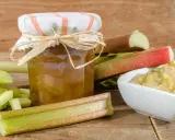 Un bocal de confiture de rhubarbe pas trop sucrée, entouré de tiges de rhubarbe fraîchement coupées et d'un bol de compote.