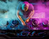 DJ con cuffie e cappellino mixa musica hause su console illuminata da luci viola e blu, circondato da fumo.