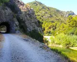 Camino de grava junto a un río y un túnel en la vía verde de Alicante. Naturaleza exuberante y rocas escarpadas.