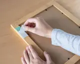 Mano retirando cinta de doble cara de un marco de madera. Aprende cómo quitar cinta de doble cara de la pared sin dañarla.