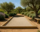 Terrain de pétanque en gravier, bordé de bois, sous des oliviers. La réglementation du jeu est respectée pour ce lieu paisible.