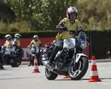 Hombre en moto blanca, con chaleco amarillo y casco, practicando maniobras en un curso carnet A. Otros motoristas y conos en el circuito.