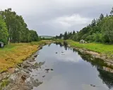 Ein ruhiger Fluss, der durch eine grüne Landschaft fließt. Kleine Häuser säumen das Ufer, vielleicht Orte, an denen man das schottische Uisge Beatha genießt.