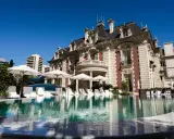 En lyxig pool framför en ståtlig byggnad som påminner om White Lotus Hotel, med solstolar och parasoller.
