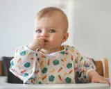 Niebieskooki maluch w śliniaczku z warzywami, kiedy dziecko rozpoznaje twarze, z paluszkiem w buzi.