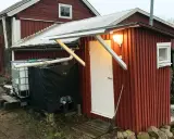 Rött utedass med vit dörr och fönster. Bredvid står en stor tank och en svart presenning. Här följer man utedass regler.