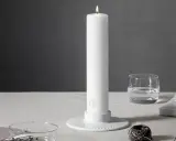 Ett vitt, högt stearinljus med guldig logotyp står i en vit ljusstake. Bredvid finns ett glas vatten, böcker och garn. Olika former av stearinljus kan skapa stämning.