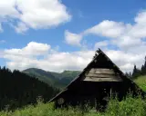 Drewniany szałas pasterski w Tatrach, otoczony zielenią i górami, pod błękitnym niebem z białymi chmurami.