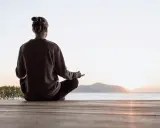Person sitter i lotusställning på en träbrygga vid havet, med solnedgång i bakgrunden. En stund av mindful yoga.
