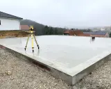Der **Bodenplatten-Aufbau** ist fast fertig. Ein Nivelliergerät auf einem Stativ steht auf der frisch gegossenen Betonplatte. Im Hintergrund sind Häuser und eine Kirche zu sehen.