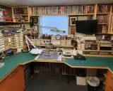 Ein Hobbyraum für **Plastikmodellbau**: Regale voller Modelle, Farben und Werkzeuge, eine Werkbank mit Schneidematte und Lupenlampe.